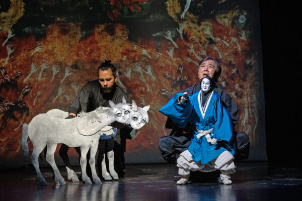 Clor photo of two puppeteers dressed in black on stage. They each manipulate figures - a puppet dressed in traditional Japanese clothing on the right and two stylized horses with human faces on the left. Behind them a brightly colored backdrop featured many reaching human hands.