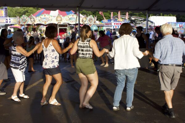 Color photo of a group of people shot from the back - hands joined in a folk dance. They wear street clothes and in the background, a carousel and ticket booth can be seen.