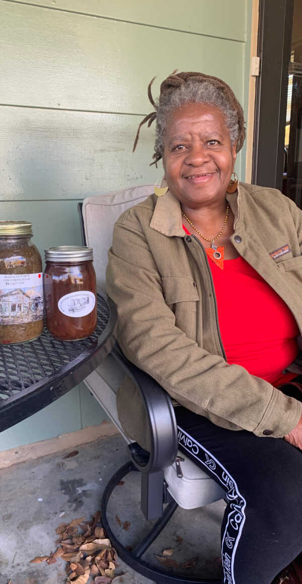 Color photo of a woman sitting on her porch with jars of preserves on a table beside her.
