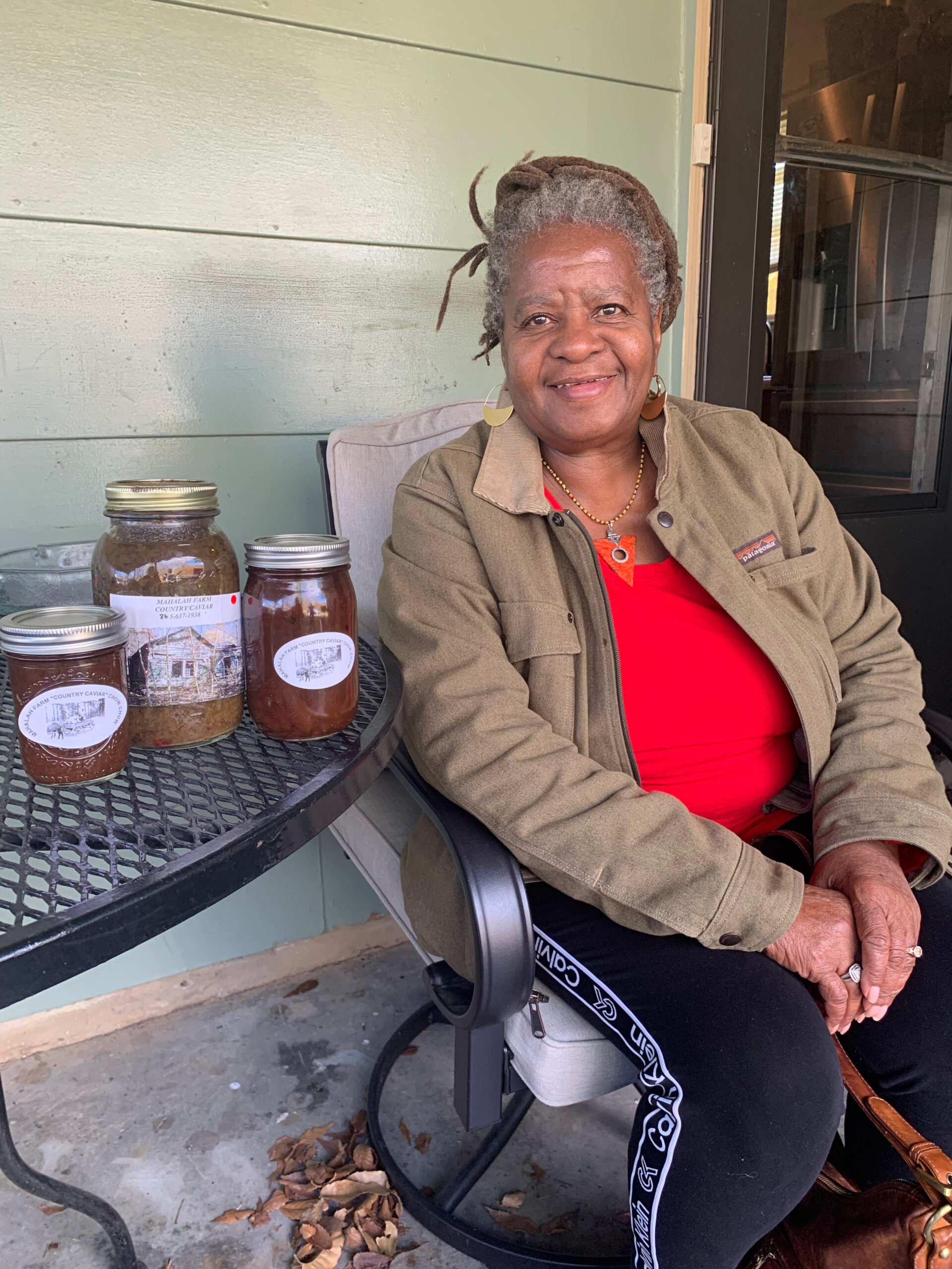 Color photo of a woman sitting on her porch with jars of preserves on a table beside her.