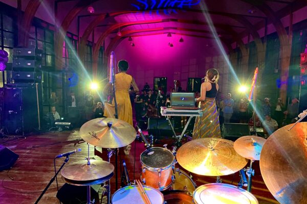 Color photo shot from behind two performers on stage with mics singing to an audience in a venue. In the foreground, a full drum kit can be seen.