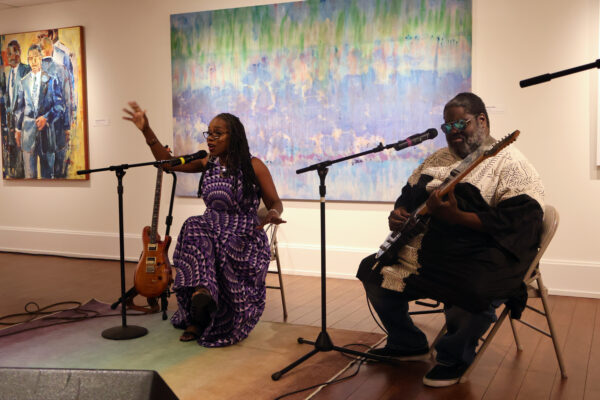 Color image of two artists seated on chairs with stand mics performing in a space with large paintings on the walls behind them.