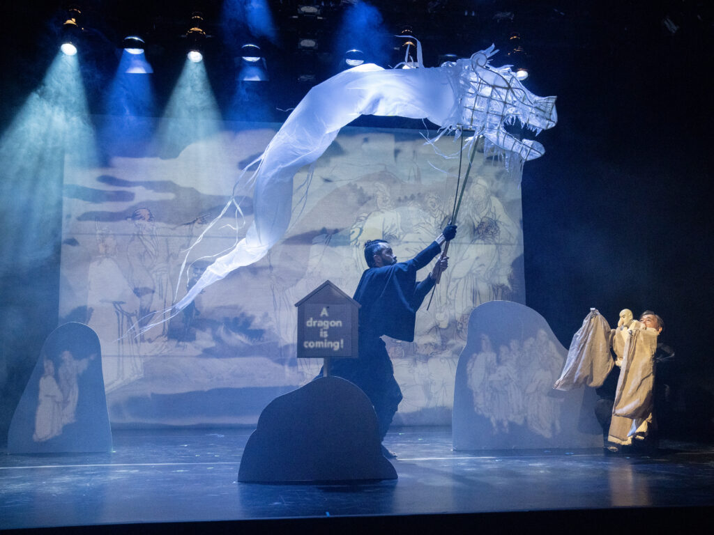 A puppeteer maneuvers a large dragon on a stick across a large stage. A figure with arms raised stands to the right.