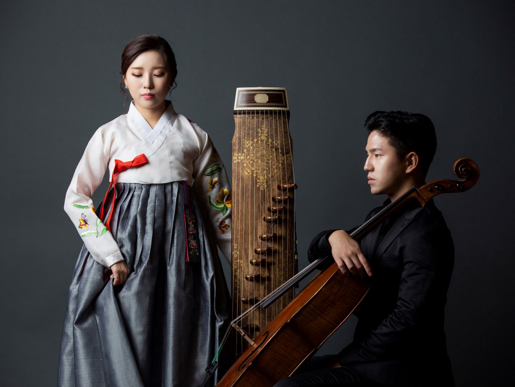 Color photo of CelloGayageum. Shot in front of a medium grey background, a woman in traditional Korean dress stands left one hand holding her long silk skirt and the other balancing a Gayageum, a traditional stringed instrument. To her right, a man sits in side profile holding a cello. He wears a dark suit.