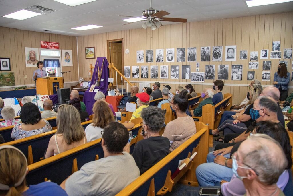 A color photo of a large group of people facing a speaker at a podium in a meeting room. The people sit on wooden pews and the walls are lined with black and white photographs.