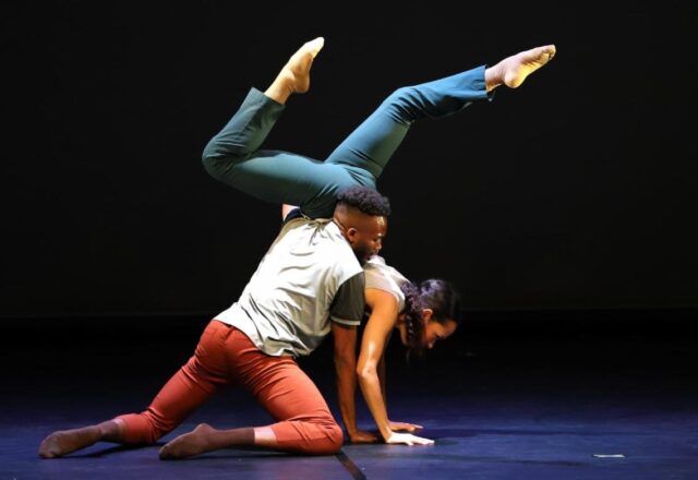 Color photo of two dancers. One kneels and supports the other in a split handstand.