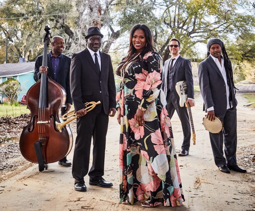 Color photo of the band Ranky Tanky. The four musicians stand on a dirt road in a v shape. A woman in a long flowered dress stands at the front flanked by four men in dark suits.