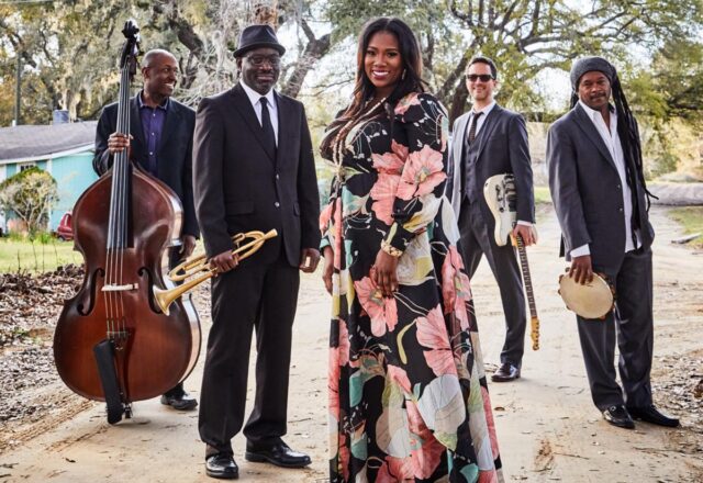 Color photo of the band Ranky Tanky. The four musicians stand on a dirt road in a v shape. A woman in a long flowered dress stands at the front flanked by four men in dark suits.