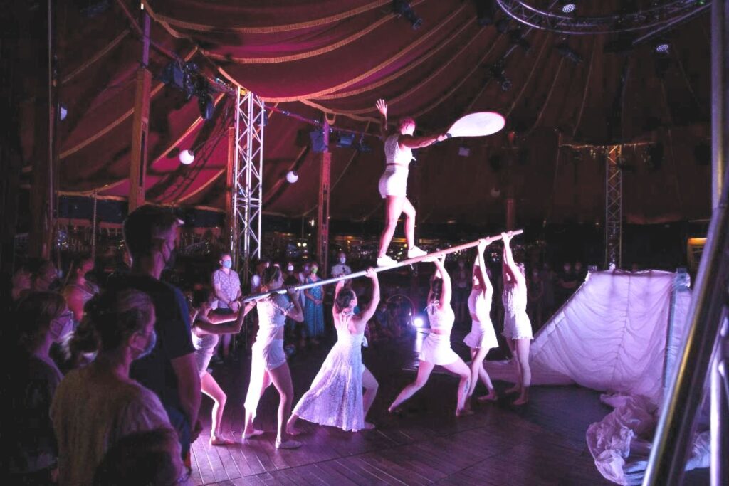 Inside a bigtop tent, a group of women all in white clothing, hold a long pole over their heads. Another woman balances on top the pole.