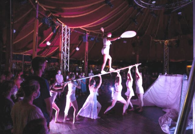 Inside a bigtop tent, a group of women all in white clothing, hold a long pole over their heads. Another woman balances on top the pole.