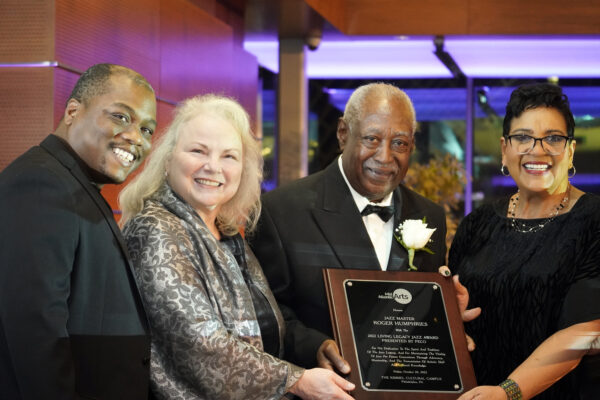 Color photo or four people on a stage looking at the camera. In the center, a gentleman holds a plaque and wears a tuxedo.