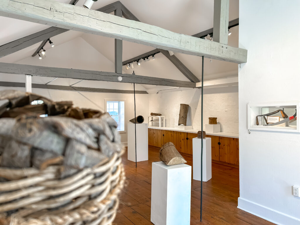 Color photo of a large open white gallery with wood beams showcasing basketry and the tools and materials used to create the pieces.