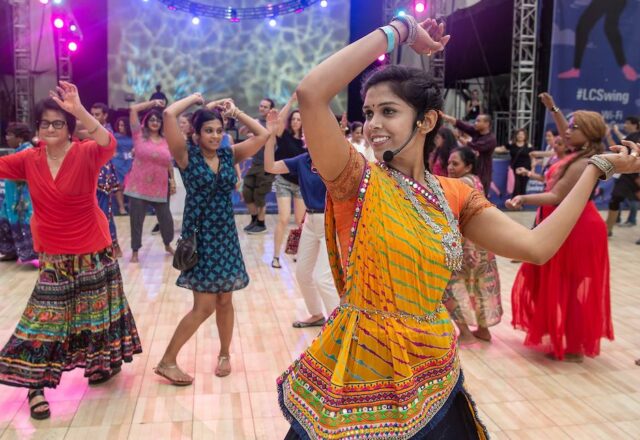 Colorful image of an artist leading community members through the steps of a dance. A large festival stage is lit in the background.