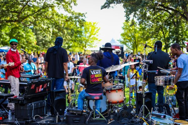 Color photo shot from behind an outdoor stage of a large jazz band facing an audience in a grove of trees.