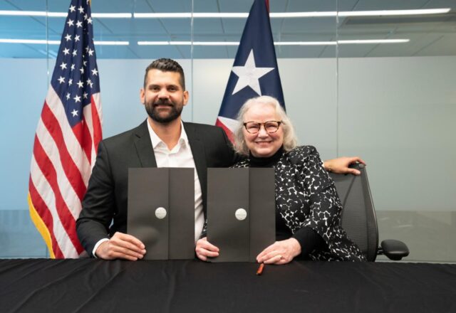 Color photo of Carlos Ruiz and Theresa Colvin at the signing of the Puerto Rico partnership agreement. In the background the US and Puerto Rican flags can be seen,