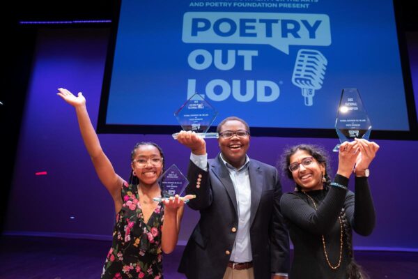 Color photo of three students standing on stage lifting glass trophies. Behind them the Poetry Out Loud logo is projected on a large screen.