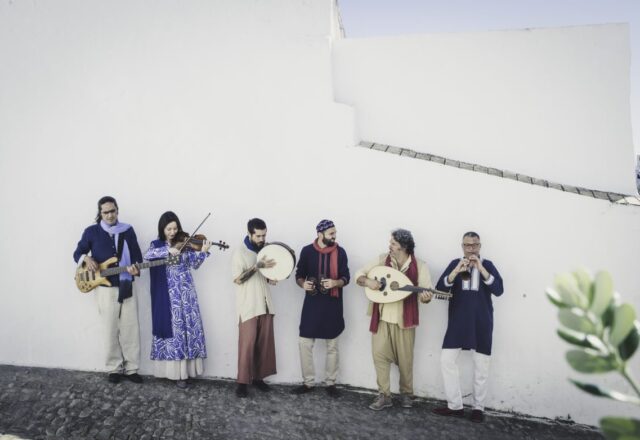 Color photo of six musicians wearing traditional garb holding their instruments against a stark white adobe wall.