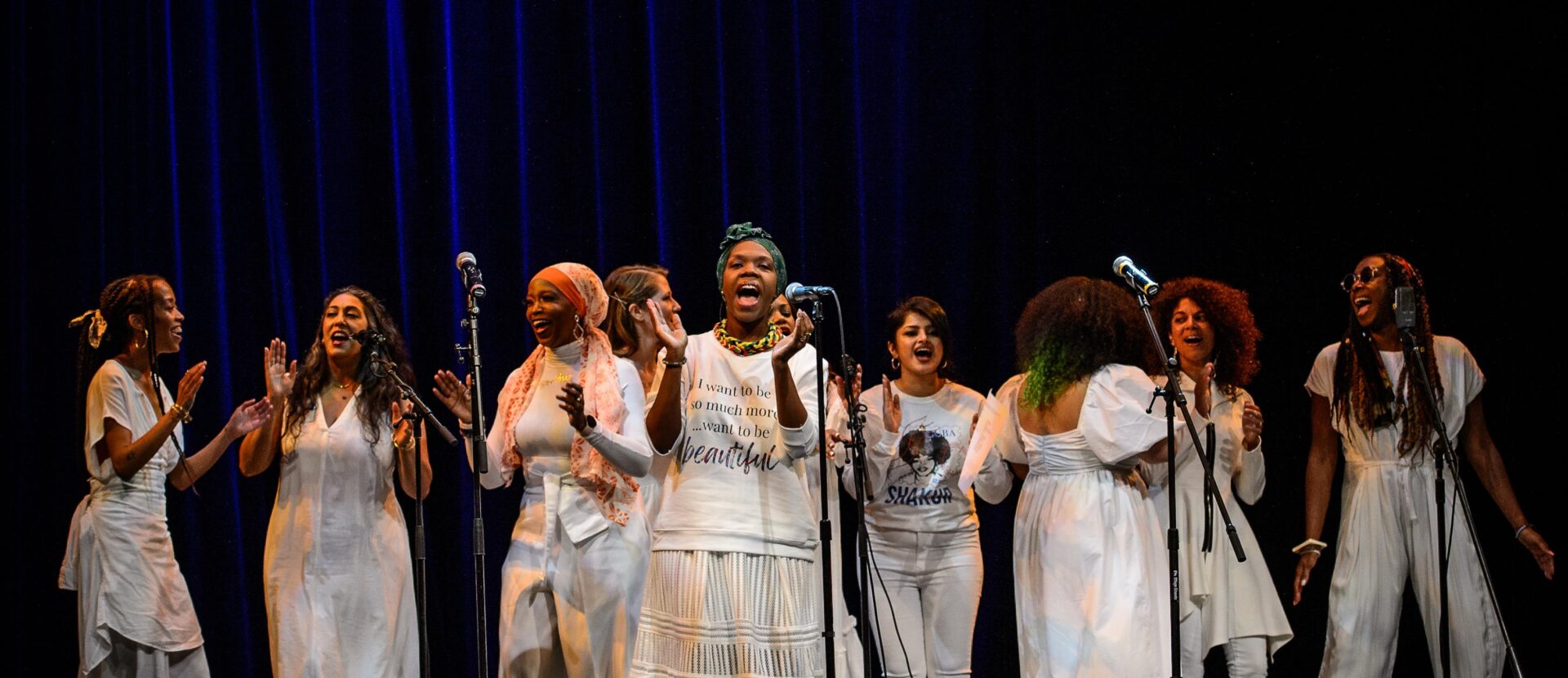 Color photo of a large group of women dressed all in white singing and clapping onstage in front of a dark blue curtain.