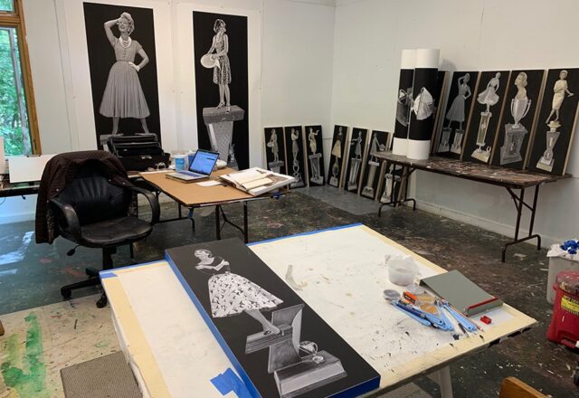 An art studio with white walls and a paint splattered floor. Two tables hold art, tools, research books and a laptop. Finished pieces line the walls.