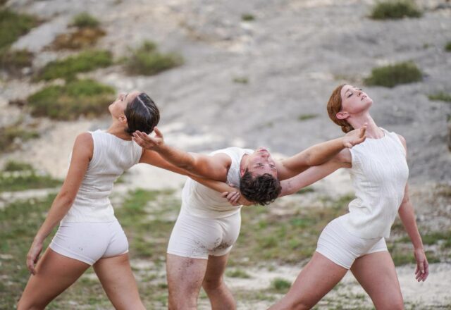 Color photo of three dancers in white shot outdoors. They strike a pose in a rocky area with short grass.