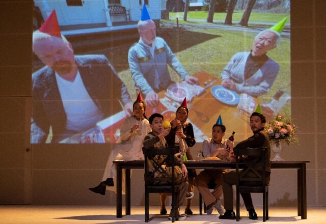 A group of five actors sit at a dining table onstage. Behind them, on a very large screen, a group of three people can be seen. Both groups are hosting a party and all of the cast members wear party hats