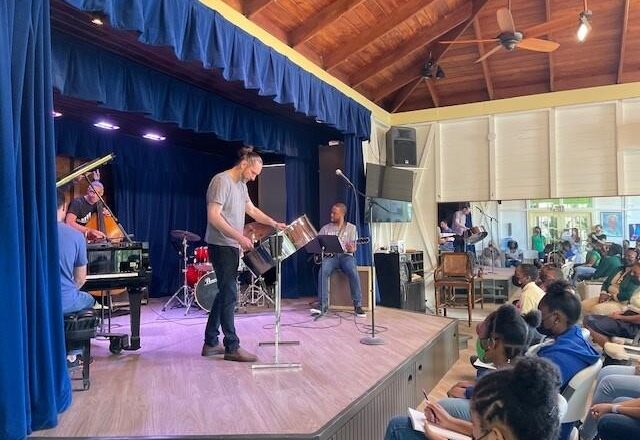 Color photo of musicians on stage in a large room with a wooden ceiling. The musician at the front plays steel pan.