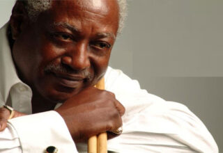 Color photo of Roger Humphries. He is a black man with short grey hair a a mustache. He holds drumsticks and wears a white shirt.