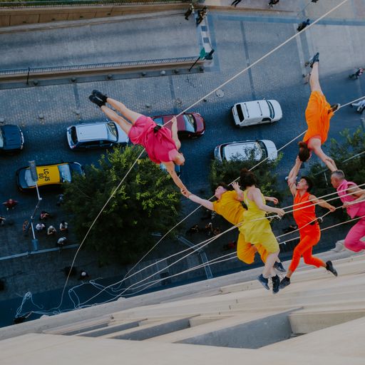 Color photo of a dance company dressed in bright color suspended by wires off the side of a building. Cars and the street can be seen below.