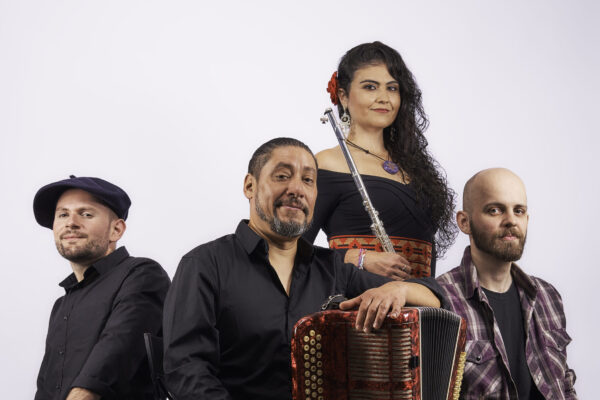 Three males musicians site on boxes in a semicircle. They all ear shades of black with accents. To the left a bass lays on the floor. The center musician holds an accordion. The man to the right has an acoustic guitar. Behind them standing center is a woman with a flute.