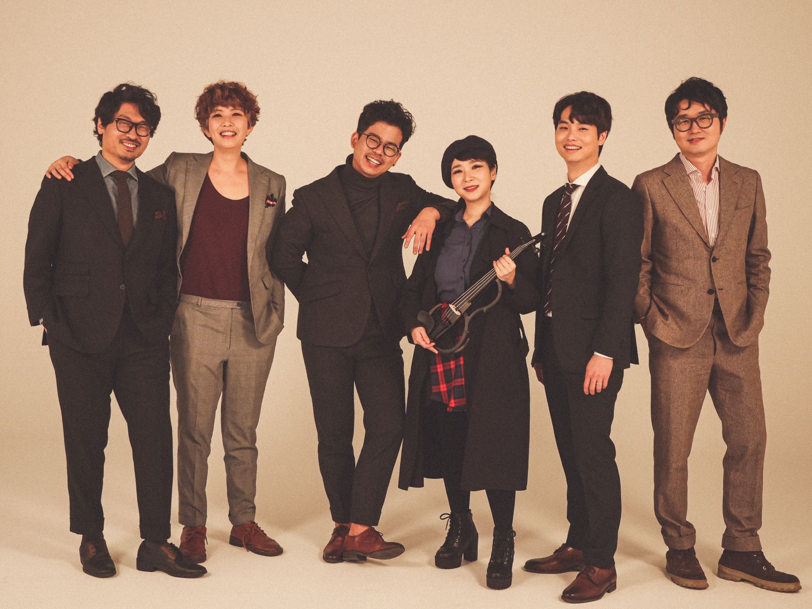 Color photo of 2nd Moon. Six people stand in front of an off-white studio background. They are dressed in a variety of suits and shirts and a woman standing center hold a stringed instrument.