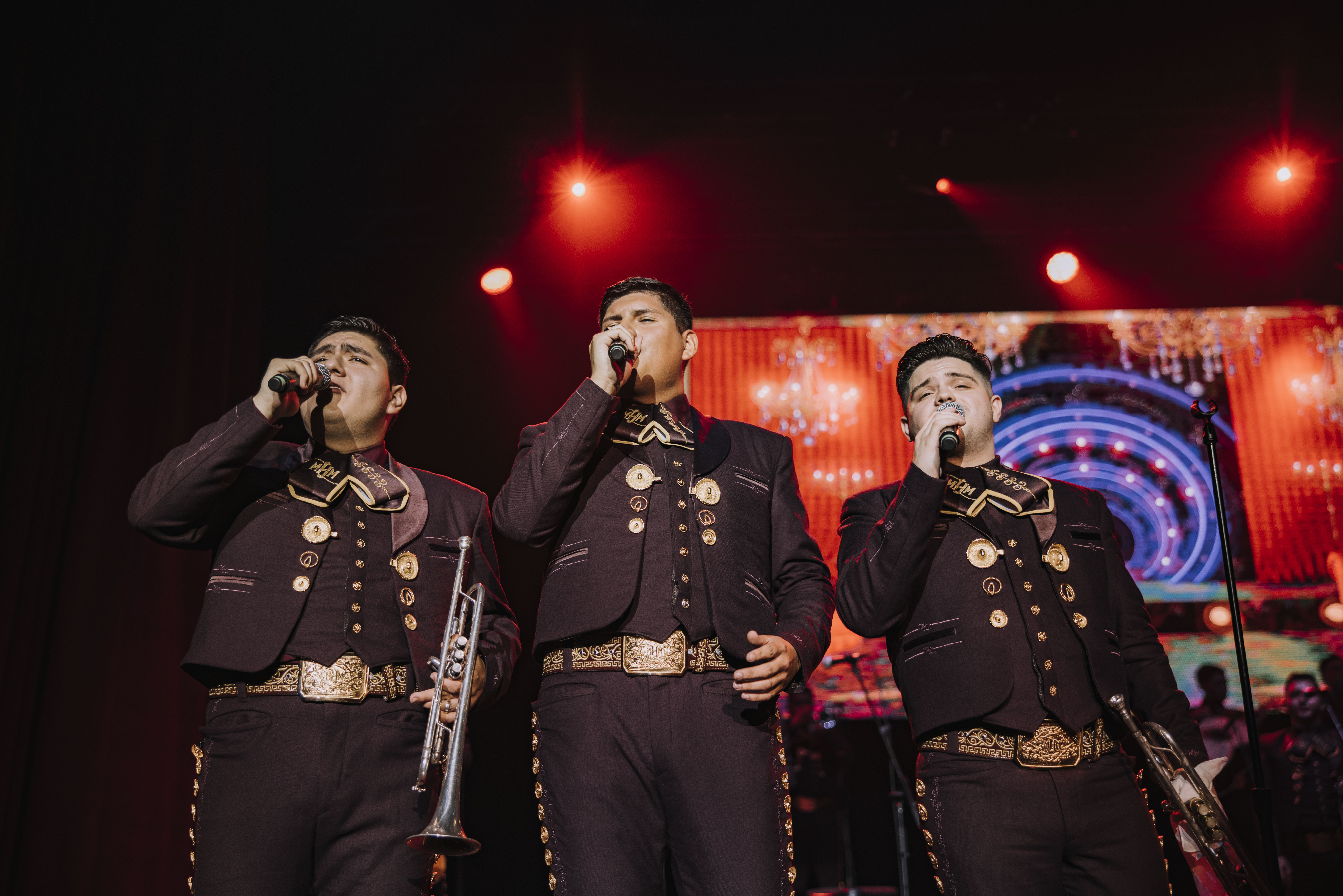 Color photo of three artists singing into hand mikes on a stage. They wear traditional costumes of the Mariachi.