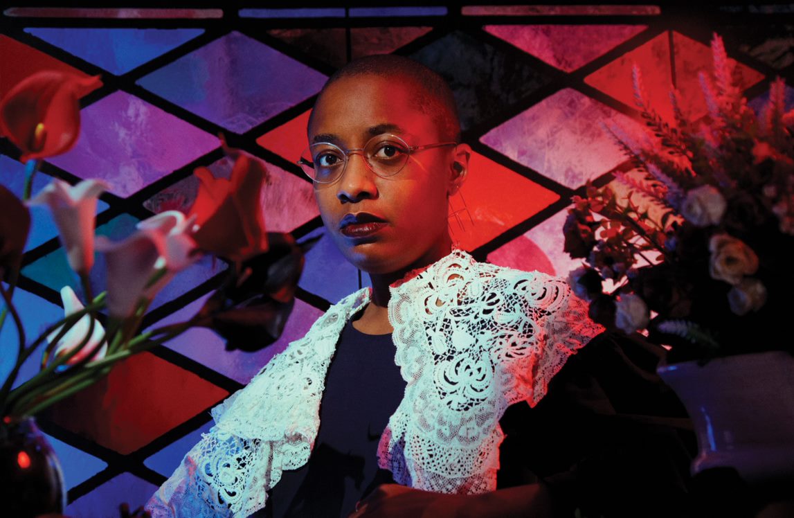 Color photo of Cecile McLorin Salvant shot against a stained glass window. Cecile wears a white lace collar.