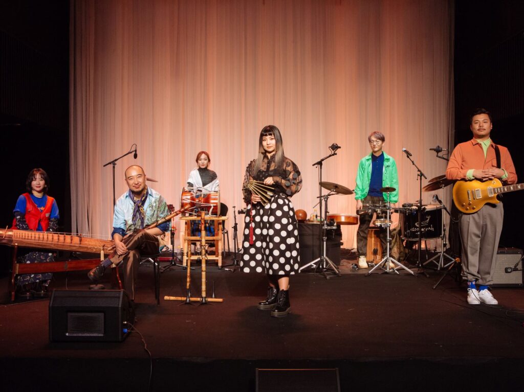 A group of musicians stands on a stage with their instruments and microphones.  This is a posed publicity photo.