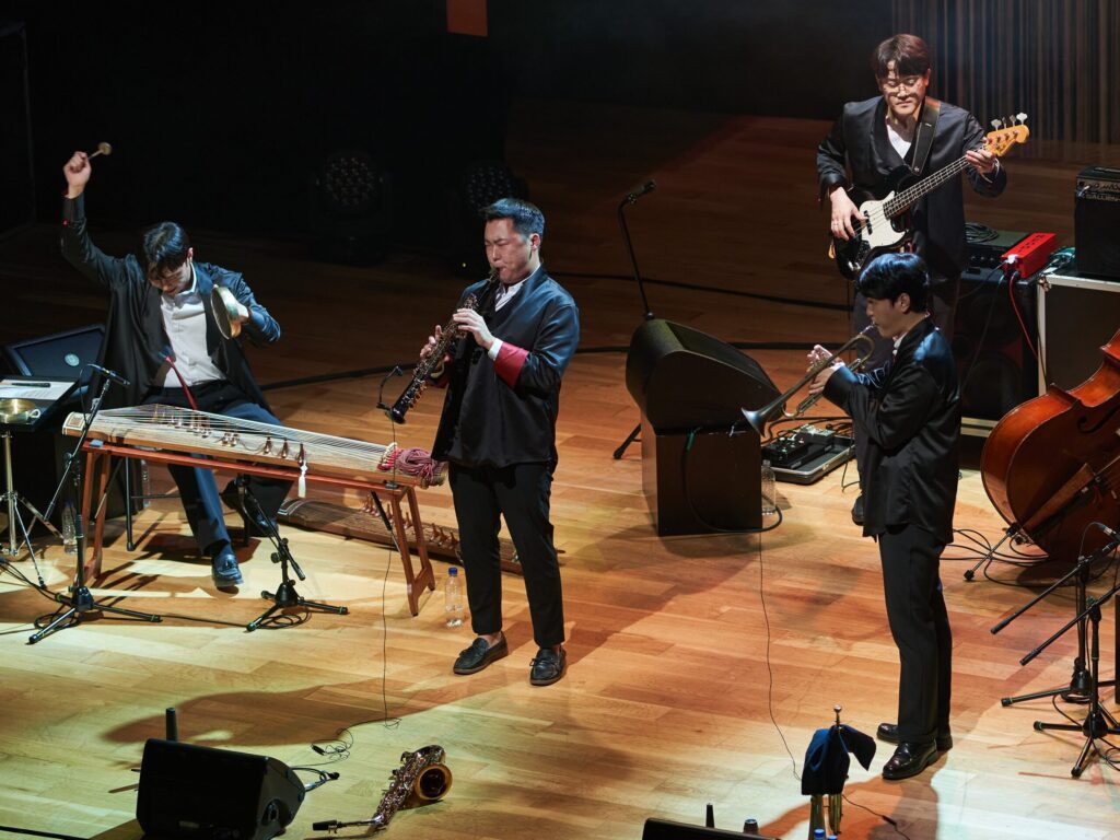 Color photo of SE;UM. Four members on the musical group SE:UM perform on a wooden stage. They are all dressed in black suits and play a variety of modern and traditional instruments.