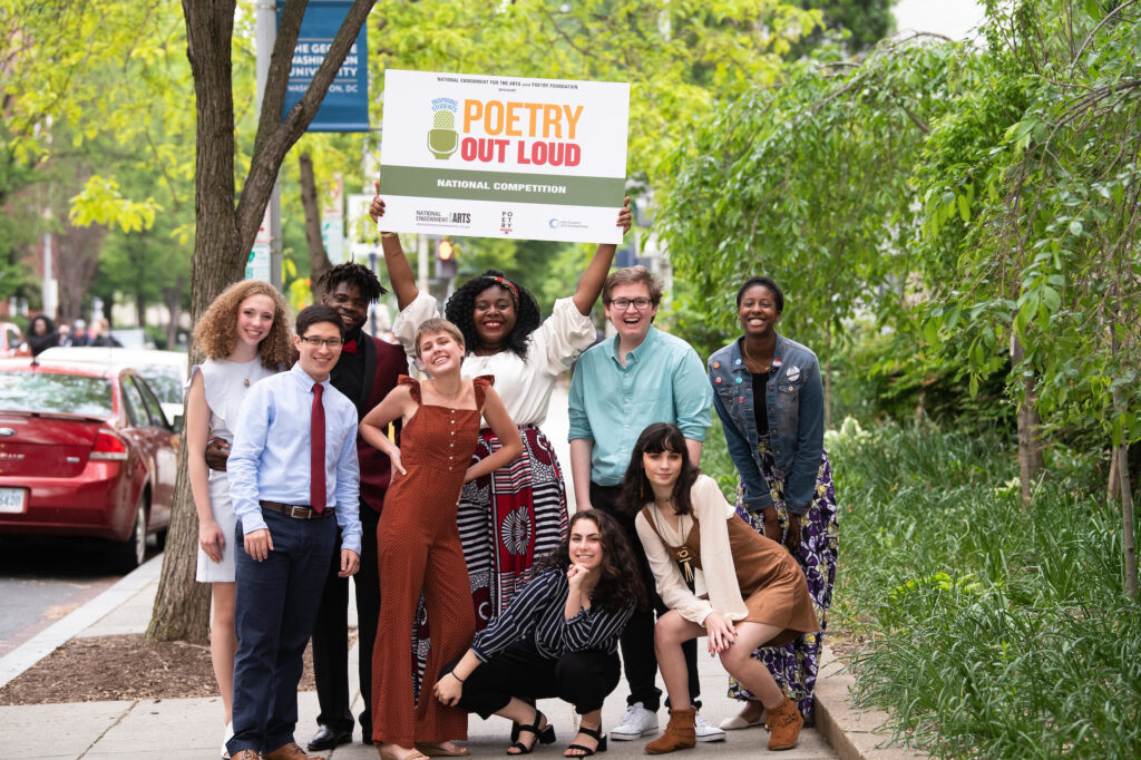 Color photo of a group of young adults posing on a sidewalk holding up a sign that says Poetry Out Loud.