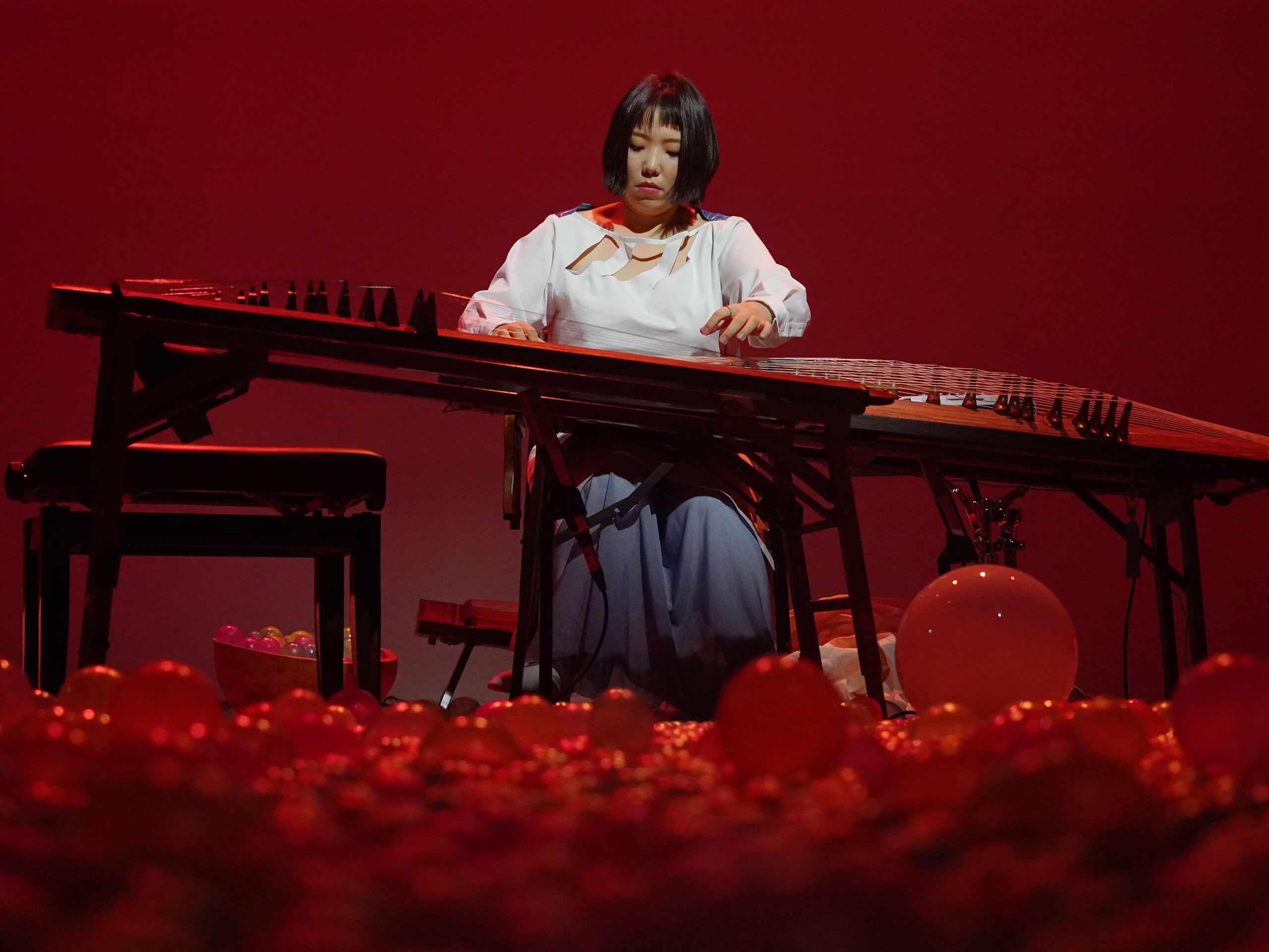 Color photo of Gayageum artist Seo Jungmin. Seo sits center playing the Gayageum. Multicolor balls of all sizes and colors cover the ground at her feet. Bathed in red light she wears a white blouse and grey slacks.