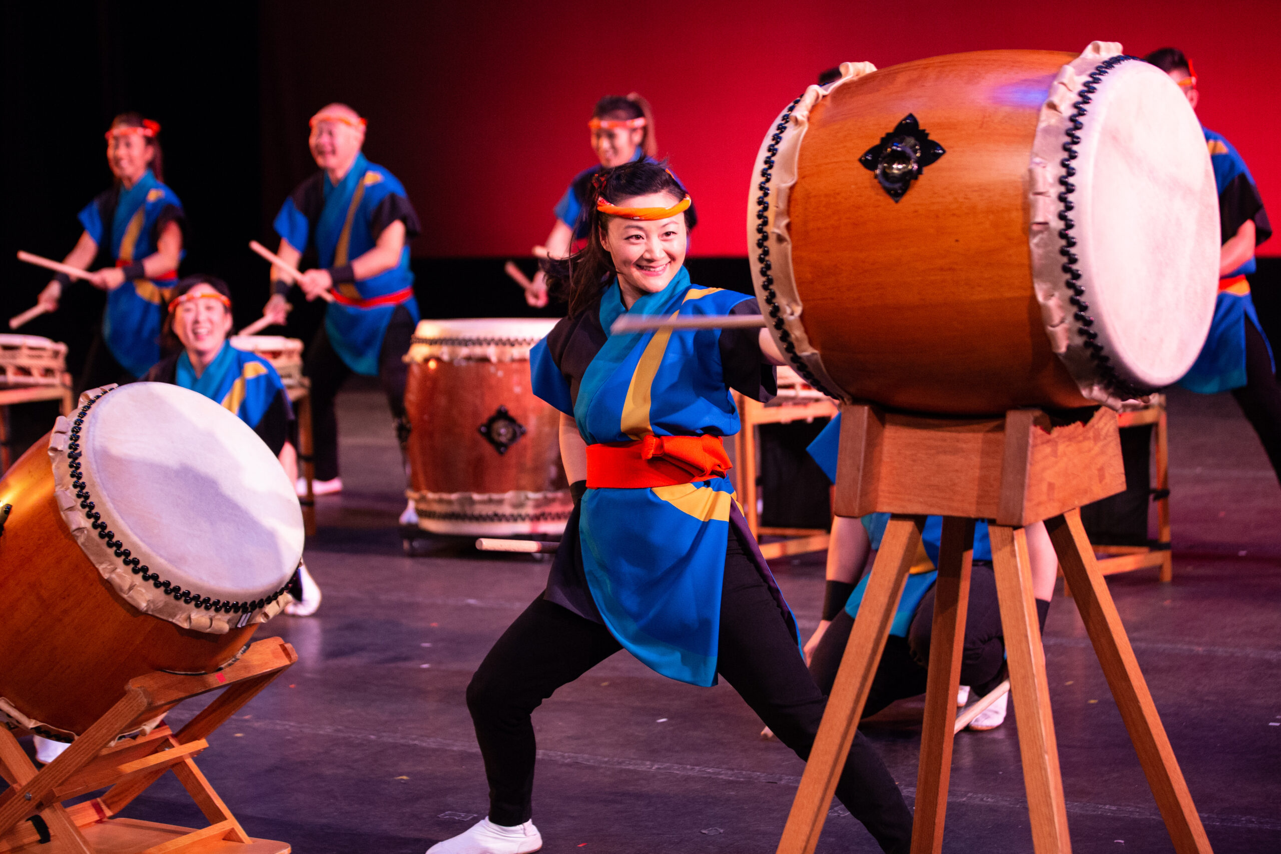 Color photo of traditionally dressed drummers playing large drums on a stage.