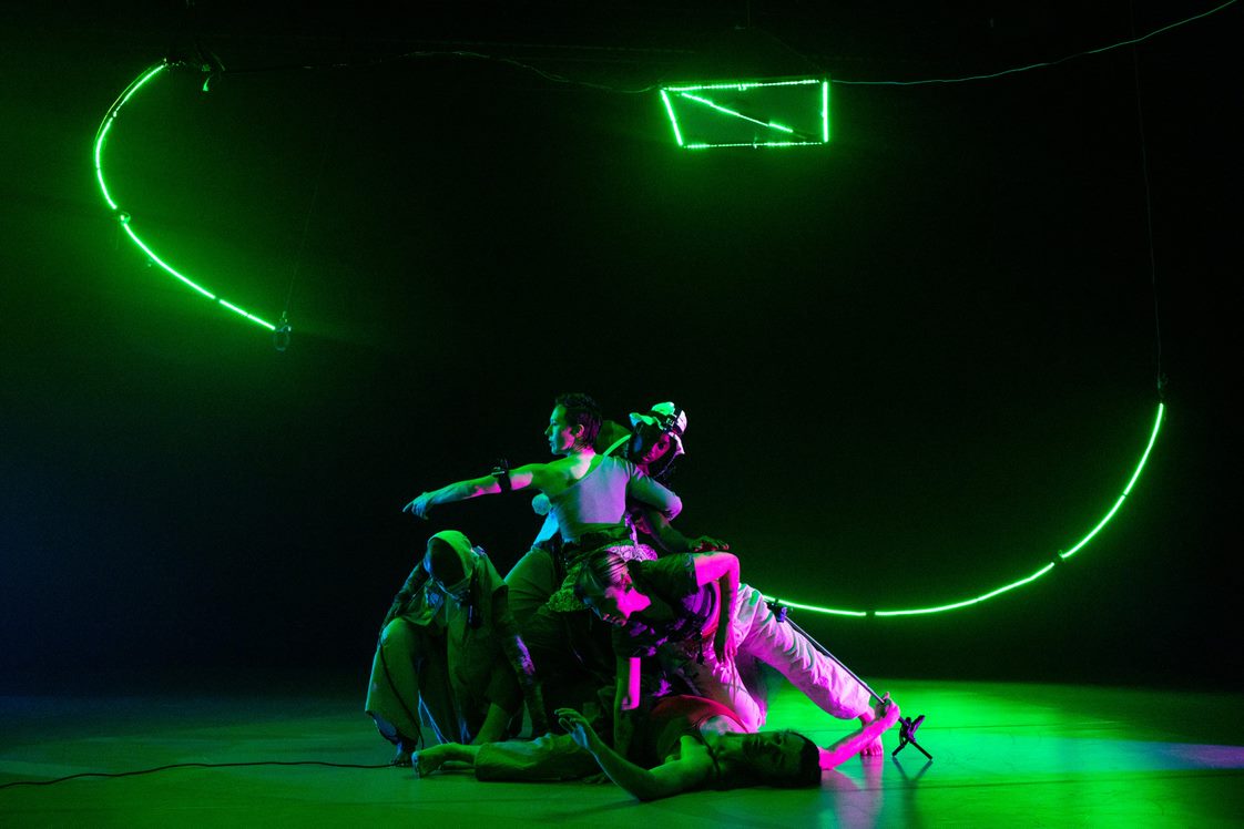 Color photo of slowdanger dance company on a stage bathed in green. Some of the dancer lay on the stage, some crouch, and some stand.