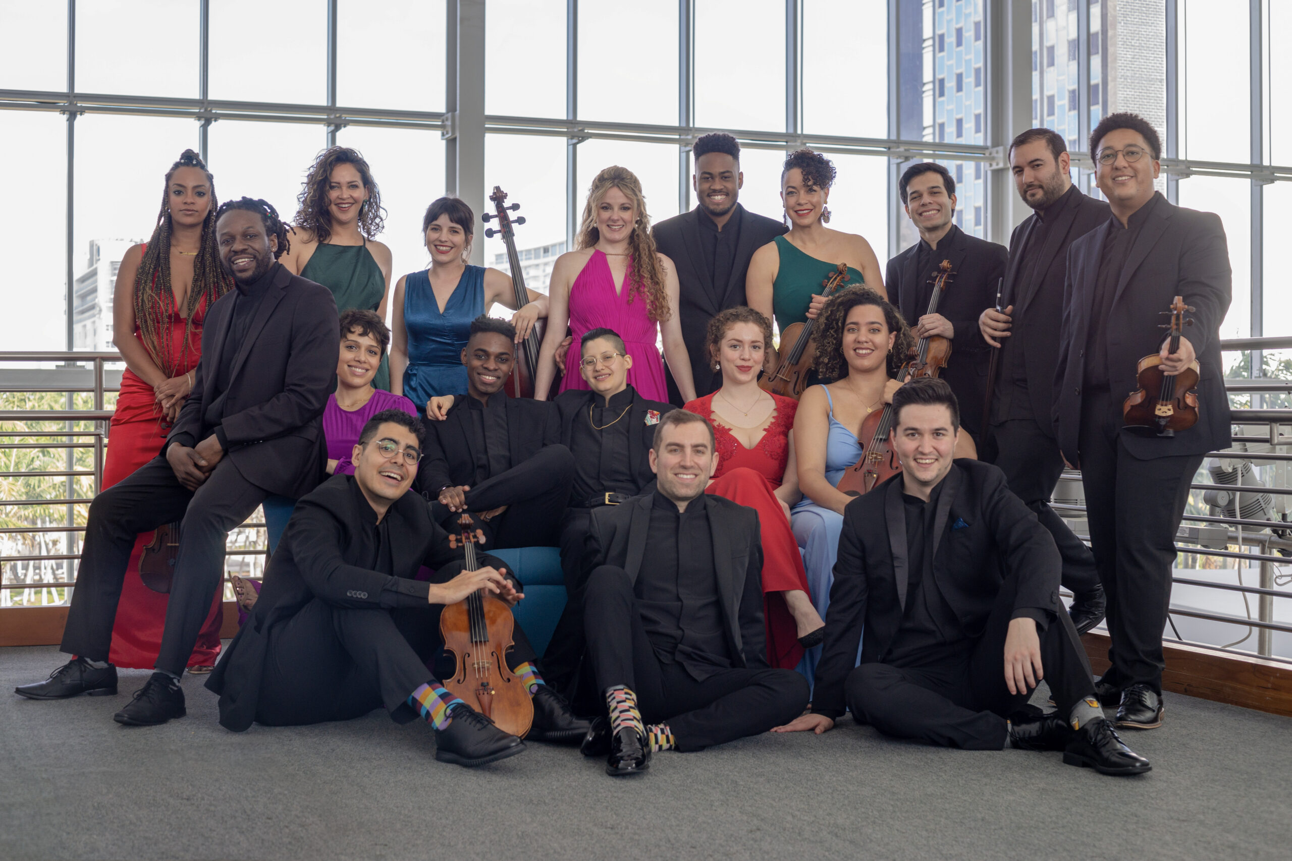 A large music ensemble poses for a group picture in front of floor to ceiling glass windows in a city.