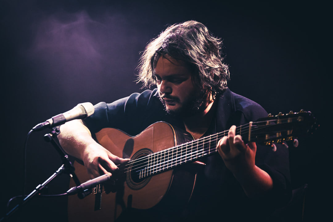 Close-up color photo of a man playing a guitar with a stand mic in front of him.