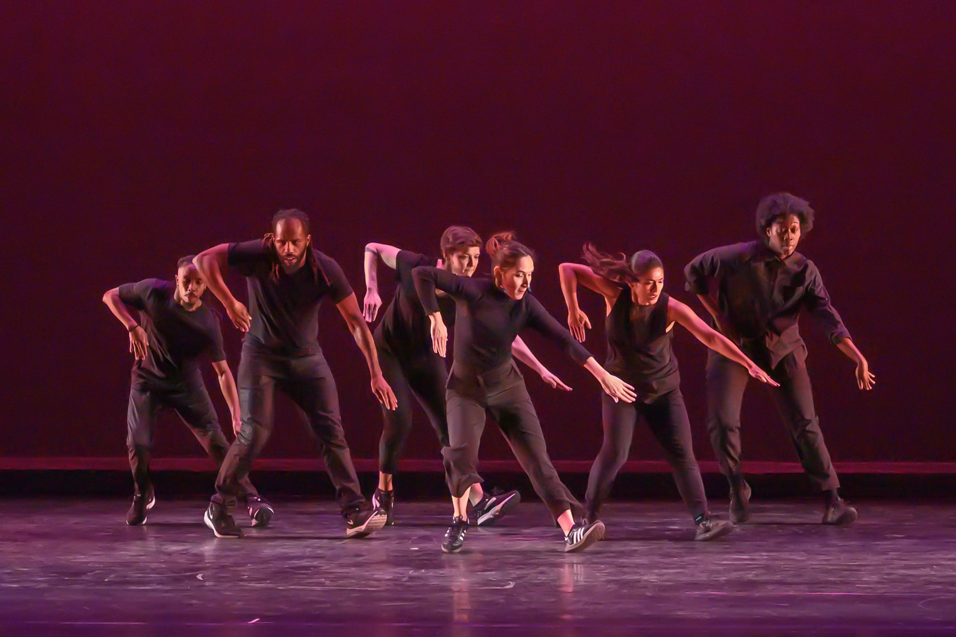Color photo of six dancers onstage all in black one arm extended toward the floor.
