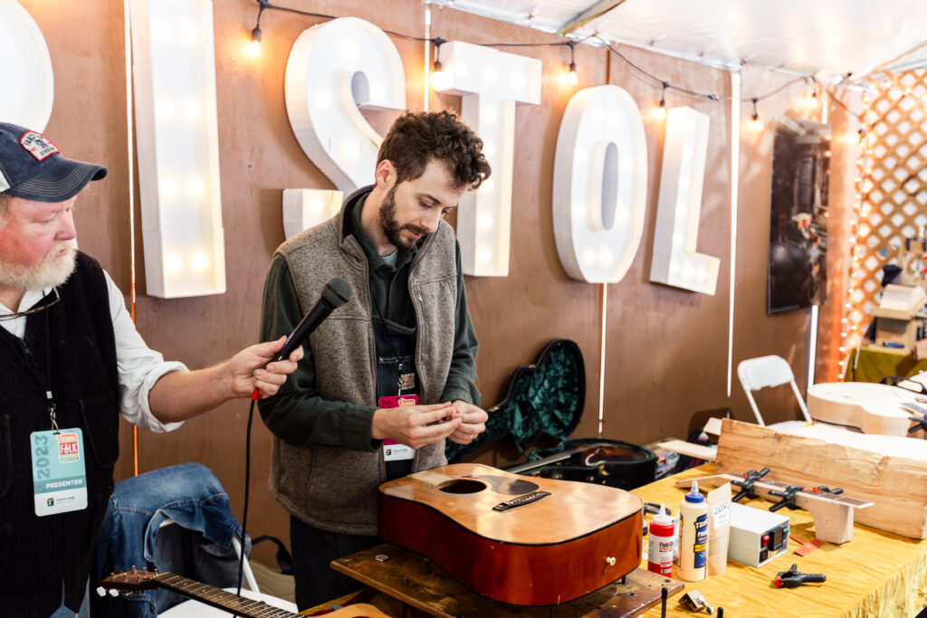 Color photo of a person repairing a guitar while someone holds a microphone for him to speak into. The word Bristol is large on the tent wall behind the demonstrator.