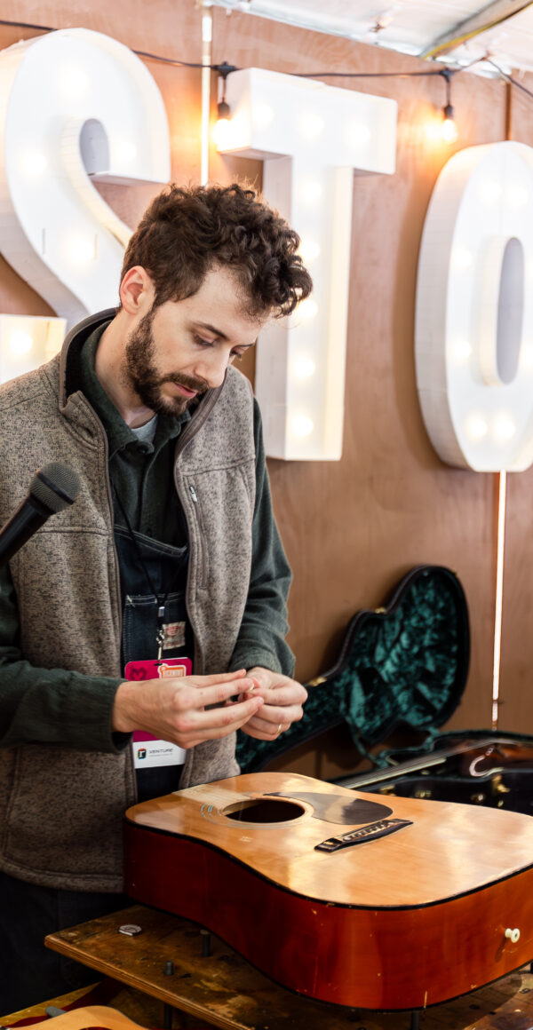 Color photo of a person repairing a guitar while someone holds a microphone for him to speak into. The word Bristol is large on the tent wall behind the demonstrator.