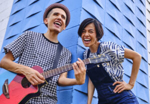 Andrés and Christina, medium-skinned Latinx performers, in center of frame, full body with arms outstretched, standing on concrete steps, with a curvy building covered in metallic tiles in the background.