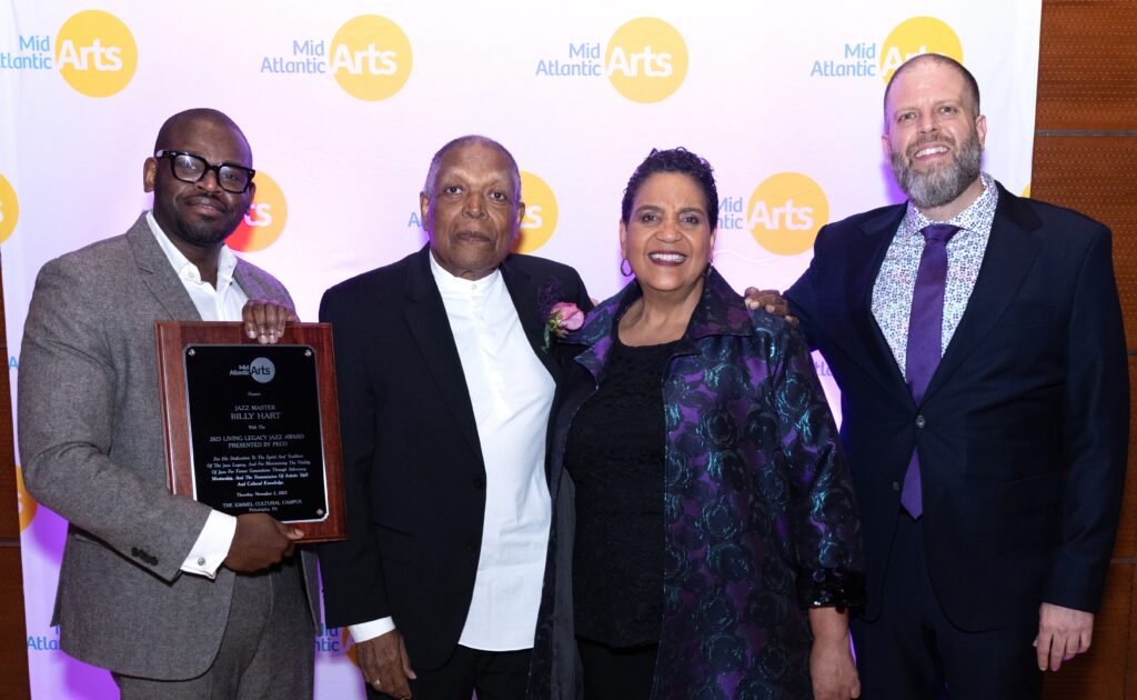 Color photo of four people standing in front of a backdrop with the Mid Atlantic Arts Logo on it. One person is holding a plaque and everyone is smiling.