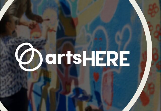 The artsHERE logo features two overlapping spheres in green and orange. It’s framed by a circular outline. The logo is over a blurred image of folks of color painting a colorful mural.