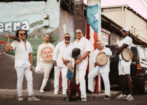 Six musicians dressed in white and holding instruments pose in front of a brightly painted street corner.