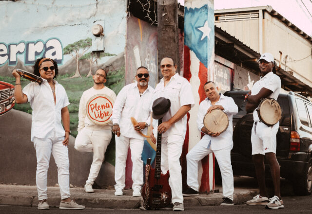 Six musicians dressed in white and holding instruments pose in front of a brightly painted street corner.