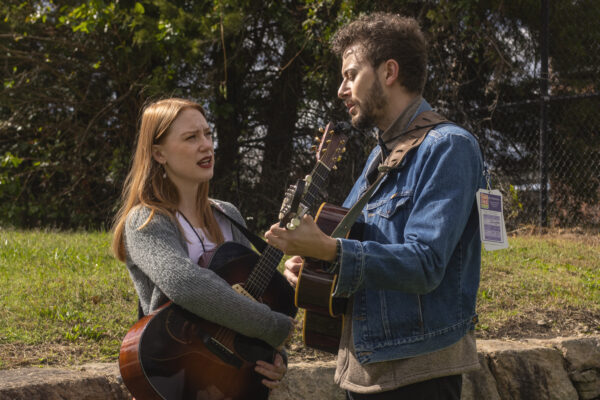 Two musicians stand outside by a stone wall and grass holding guitars and signing.