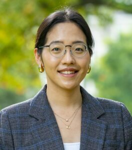 Kelly Jung is a woman with a medium-light skin tone, dark hair pulled up, and wire rimmed glasses. She wears a white top and grey plaid blazer.